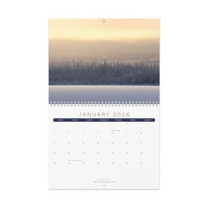 May include: A wall calendar for January 2026, featuring a winter landscape photograph. The image shows a snow-covered lake and trees under a hazy sky. The calendar has a spiral binding and a simple layout with dark blue headers.