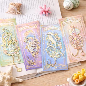 May include: Four decorative metal pins with mermaid and sea creature designs. Each pin is on a card with a pastel color scheme and gold accents. The pins feature intricate details and dangling chains with charms. The cards have text at the top.