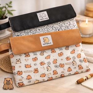 May include: Two rectangular pouches, one black and one tan, with flap closures. The tan pouch features a repeating pattern of cartoon capybaras. The black pouch has a white pattern of cartoon dogs. Both pouches have labels that read "Kappy Dog" and "Kapibala".