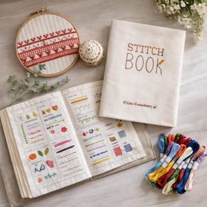 Puede incluir: Un libro de puntadas abierto con ejemplos de bordado, un libro cerrado con "STITCH BOOK" bordado en la portada y un bastidor de bordado de madera con un diseño rojo y blanco. También hay una colección de hilo de bordar colorido.