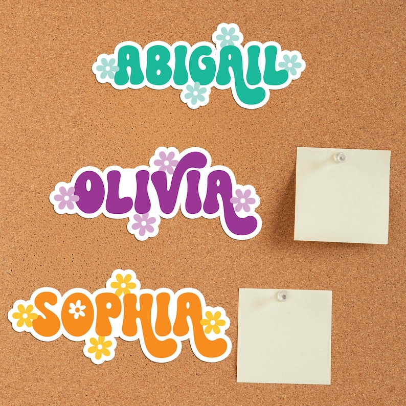 May include: Three name stickers: "Abigail" in teal, "Olivia" in purple, and "Sophia" in orange, each with a white border and flower accents. The stickers are on a corkboard, with two blank sticky notes.