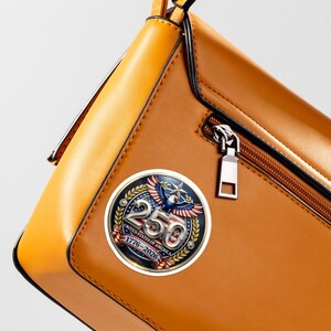 May include: A brown leather handbag with a decorative round emblem. The emblem features a patriotic design with an eagle, the number 250, and the text "UNITED STATES OF AMERICA 1776-2026". The bag has a zipper and a yellow handle.
