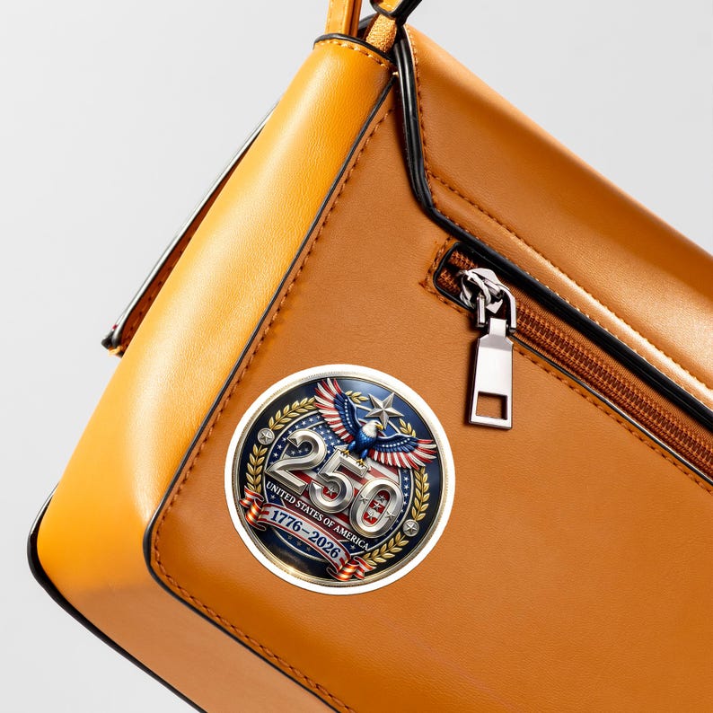 May include: A brown leather handbag with a decorative round emblem. The emblem features a patriotic design with an eagle, the number 250, and the text "UNITED STATES OF AMERICA 1776-2026". The bag has a zipper and a yellow handle.