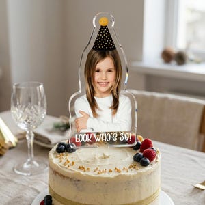 May include: A clear acrylic cake topper featuring a printed photograph of a person wearing a black party hat with gold dots. The topper is shaped like a party hat and includes the white text "LOOK WHO'S 30!" printed at the bottom base.