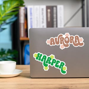 May include: Two stickers with names on a laptop. The top sticker says "AURORA" in brown letters with pink flowers. The bottom sticker says "HARPER" in green letters with yellow flowers. The laptop is on a wooden surface.