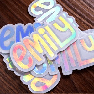 May include: A collection of die-cut stickers featuring the name Emily in a bold, lowercase sans-serif font. Each sticker has a holographic finish that reflects various colors, including yellow, pink, and blue, with a white border outline.