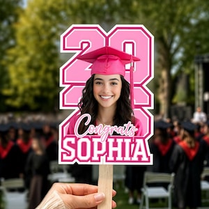 May include: A pink graduation photo prop featuring a smiling graduate wearing a cap and gown. The prop includes the year "20" and the name "Sophia" with the word "Congrats". The prop is held by a wooden stick.
