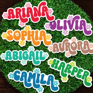 May include: Colorful name stickers with a retro font style. Names include Ariana, Olivia, Sophia, Aurora, Abigail, Camila, and Harper. Each name is in a different color and decorated with small flower accents, set against a green background.