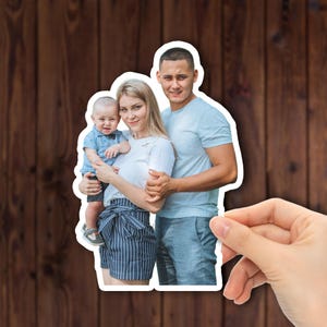 May include: A family portrait sticker featuring a smiling couple and a baby. The sticker has a white border and is held by a hand. The family is dressed in casual clothing, with the baby wearing a bow tie.