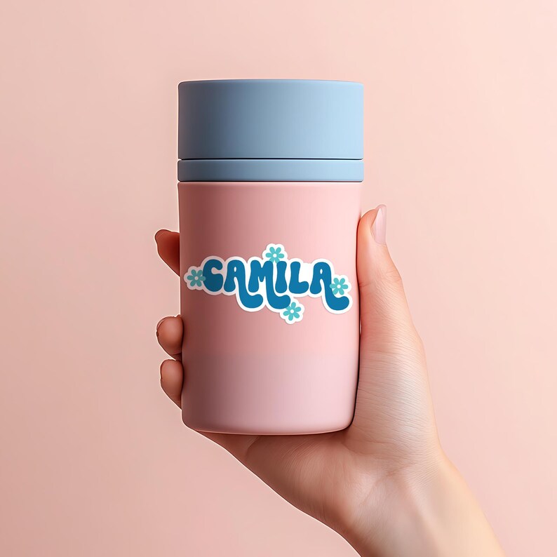 May include: A pink tumbler with a light blue lid. The word "CAMILA" is written in blue, stylized text with small blue flower accents. The tumbler is held in a hand against a pink background.