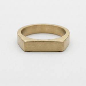 May include: A gold ring with a rectangular face and a smooth, matte finish. The ring has a simple, modern design. The ring's band is approximately 0.6 cm wide. The ring is suitable for everyday wear.