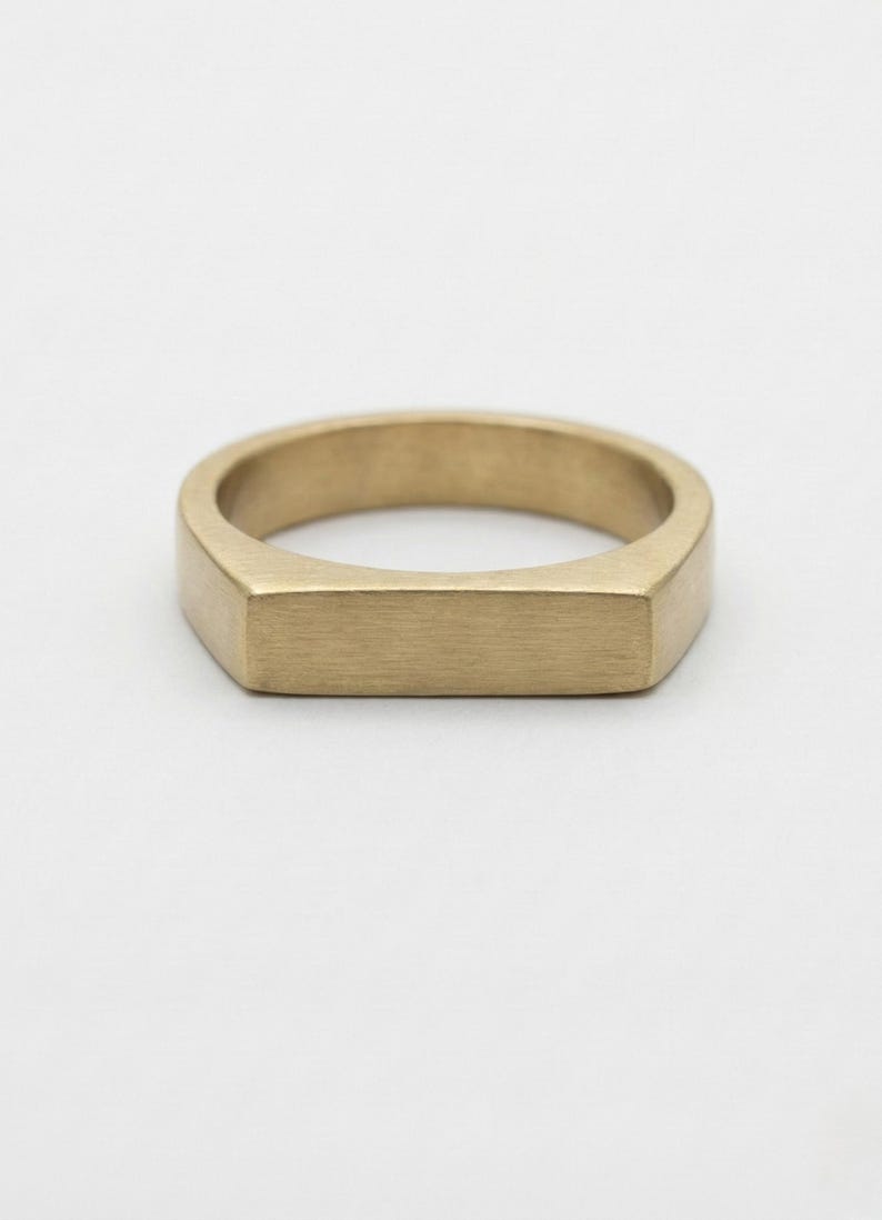 Puede incluir: Un anillo dorado con una cara rectangular y un acabado mate liso. El anillo tiene un dise&ntilde;o simple y moderno, adecuado para el uso diario. La banda del anillo mide aproximadamente 0,6 cm de ancho.