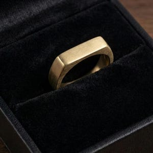 Puede incluir: Un anillo dorado con una parte superior rectangular, colocado en una caja de joyer&iacute;a de terciopelo negro. El anillo tiene un acabado de metal cepillado y un dise&ntilde;o simple y moderno. El anillo es una joya.