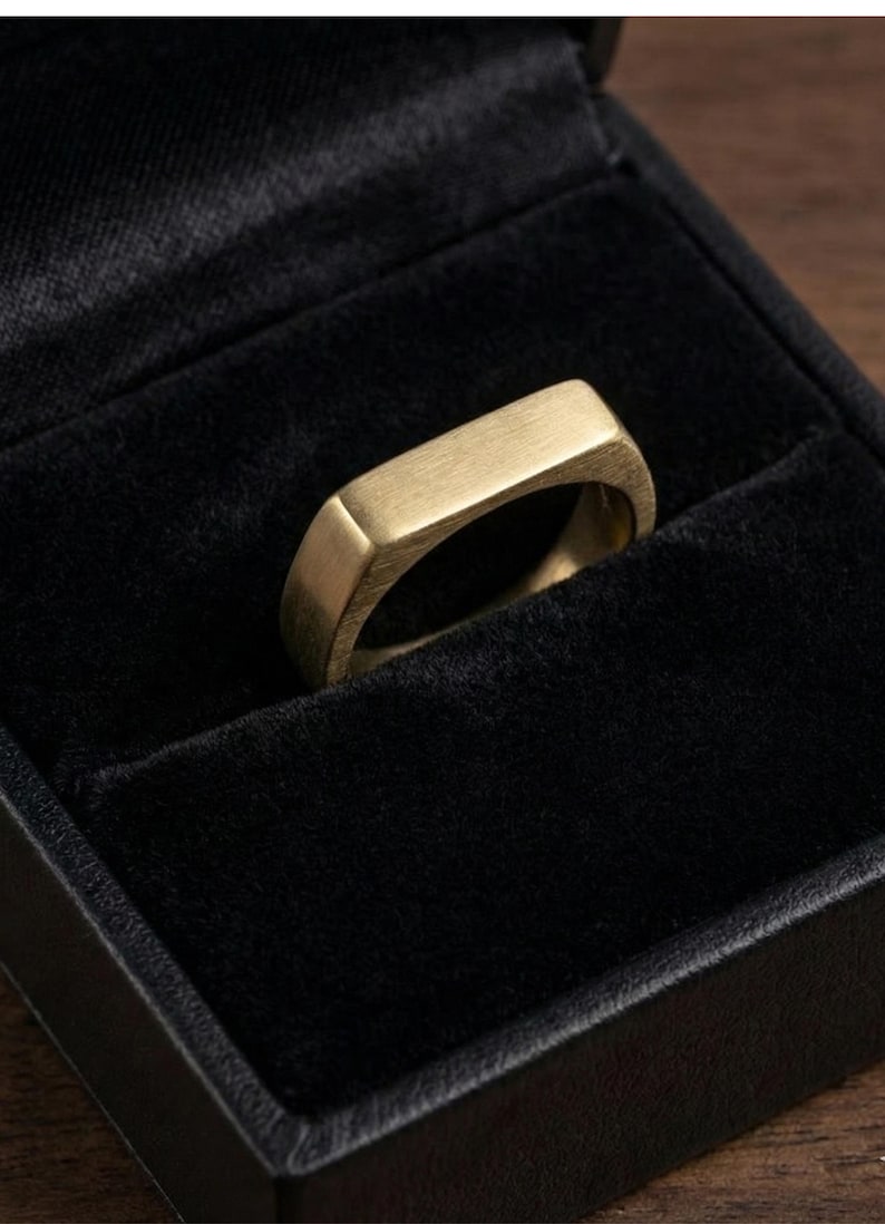 Puede incluir: Un anillo dorado con una parte superior rectangular, colocado en una caja de joyer&iacute;a de terciopelo negro. El anillo tiene un acabado de metal cepillado y un dise&ntilde;o simple y moderno. El anillo es una joya.