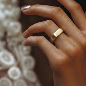 Puede incluir: Un anillo dorado con un dise&ntilde;o rectangular. El anillo se lleva en un dedo, y la imagen es un primer plano. El anillo tiene un acabado liso y mate. El fondo est&aacute; borroso, con enfoque en el anillo y la mano.