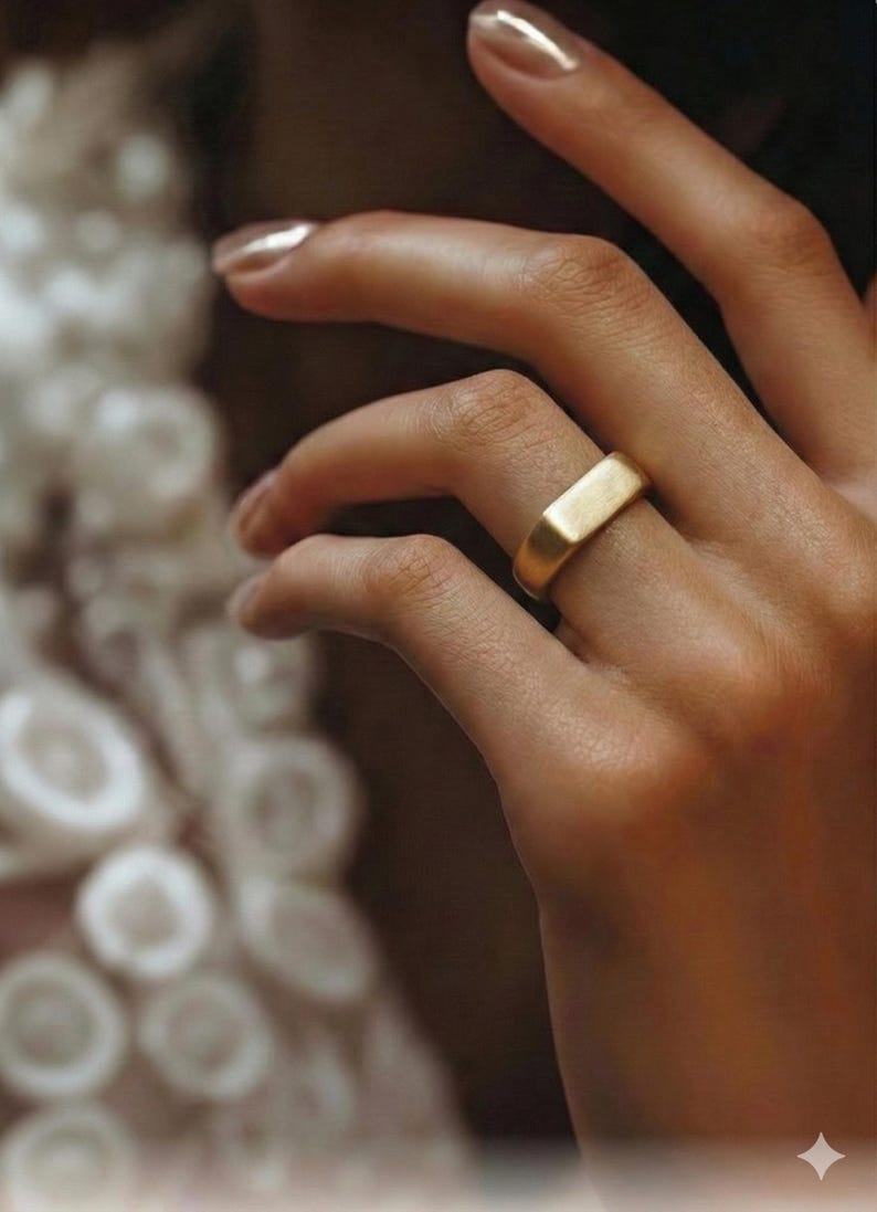 Puede incluir: Un anillo dorado con un dise&ntilde;o rectangular. El anillo se lleva en un dedo, y la imagen es un primer plano. El anillo tiene un acabado liso y mate. El fondo est&aacute; borroso, con enfoque en el anillo y la mano.