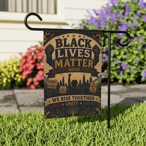 Puede incluir: Una bandera de jardín con el texto "BLACK LIVES MATTER" en letras doradas. La bandera también incluye las frases "EQUALITY, JUSTICE, FREEDOM" y "WE RISE TOGETHER". La bandera se muestra en un soporte de metal negro en un jardín.