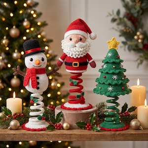 May include: Three crocheted Christmas ornaments on springs: a white snowman with a black hat and red scarf, a Santa Claus with a red hat and white beard, and a green Christmas tree topped with a yellow star, all featuring festive details.
