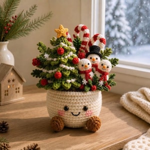 May include: A crocheted Christmas arrangement in a cream-colored pot with a smiling face. The display features a green tree with a yellow star, red ornaments, white garland, two candy canes, and three small snowmen wearing red scarves.
