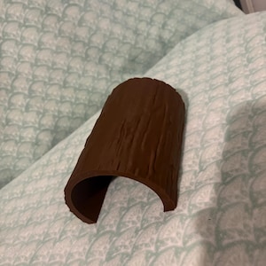 May include: A brown, semi-cylindrical object with a textured surface that mimics the appearance of tree bark. The item is curved, forming an open tunnel or arch shape, and features vertical ridges and grooves across its exterior surface.