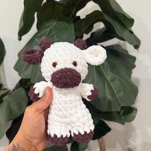 May include: A handmade crochet cow plushie. The cow is white with brown accents on the ears, muzzle, and hooves. It has small orange eyes and is held in front of a leafy green plant.