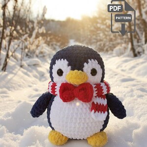 Puede incluir: Un pingüino tejido a ganchillo con cabeza y alas azul marino, cuerpo blanco, pies amarillos y pajarita y bufanda rojas. El pingüino está sobre un fondo nevado con un icono de descarga de patrón PDF.