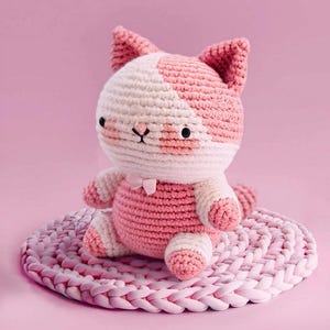 May include: A handmade crochet cat doll in pink and white, sitting on a matching pink crocheted mat. The cat has a pink bow and black eyes. The cat's body is pink, with white accents on the head, paws, and tail.