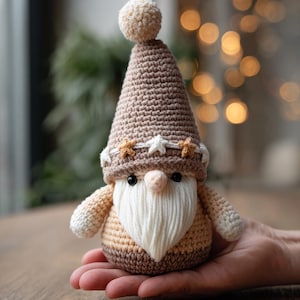 Puede incluir: Muñeca de gnomo de ganchillo con un gorro puntiagudo marrón rematado con un pompón color crema. El gnomo tiene barba blanca, nariz rosada y viste un atuendo beige y marrón. La muñeca está en la mano de una persona.