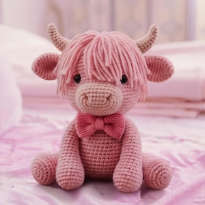 Op de afbeelding: Een handgemaakt roze gehaakt Highland-koeienpluche speelgoed met een roze strik. De koe heeft lichtbruine hoorns en oren en een franje van roze garen voor haar. Het speelgoed staat op een roze ondergrond.