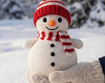 May include: A crocheted snowman with a red and white striped hat and scarf. The snowman has a carrot nose, black button eyes, and a smiling mouth. It is held in a beige mitten against a snowy background.