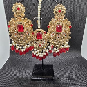 May include: A set of gold-toned jewelry featuring two large ornate earrings and a central maang tikka. Each piece is decorated with rectangular red stones, clear crystals, and small white faux pearls, with red teardrop beads hanging from the base.