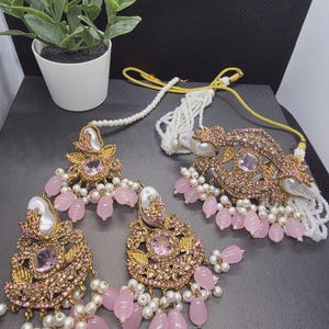 May include: A jewelry set consisting of a necklace and two matching earrings. The pieces feature gold-tone metal, light pink faceted stones, white faux pearls, and small pink beads. The necklace has a multi-strand white pearl cord with gold accents.