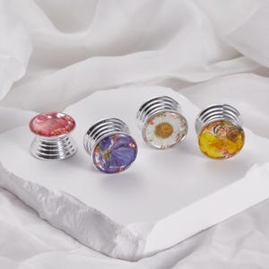 May include: Four silver-toned, decorative plugs with resin tops. Each plug features a different floral design: red, purple, white, and yellow. The plugs are displayed on a white, textured surface against a white backdrop.