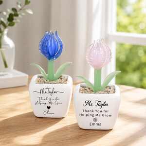 May include: Two white ceramic square planters containing glass flowers. The left planter has a blue glass tulip and text reading Ms. Taylor Thank You for Helping Me Grow with a heart and Olivia. The right has a pink glass tulip and the same text with Emma.