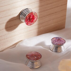 May include: Three decorative cabinet knobs with floral designs. The knobs have a silver base and a clear, rounded top filled with red and gold flowers. One knob is attached to a wooden surface, while the other two rest on a white fabric.