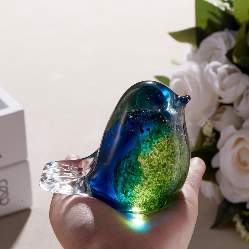 K&ouml;nnte beinhalten: Glasvogel-Figur mit blau-gr&uuml;ner Farbgebung. Der Vogelk&ouml;rper ist eine Mischung aus transparenten Gr&uuml;n- und Blaut&ouml;nen, mit einem klaren Schnabel und Schwanz. Die Figur wird in einer Hand gehalten, mit wei&szlig;en Blumen im Hintergrund.