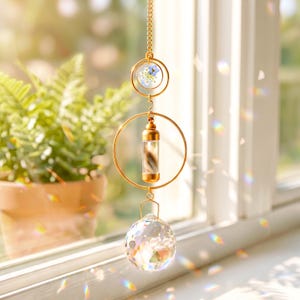 Pode incluir: Um enfeite pendurado dourado com uma bola de cristal transparente na parte inferior. O enfeite tem um pequeno frasco de vidro contendo uma pena e uma moldura circular com uma flor. O enfeite pende de uma corrente.