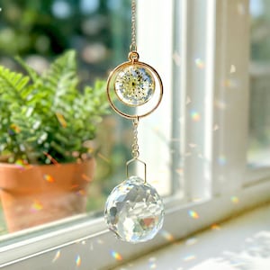 Puede incluir: Un atrapasoles colgante con un marco de metal dorado. El atrapasoles presenta una bola de cristal transparente en la parte inferior y un marco circular con flores secas encapsuladas en resina transparente. Suspendido por una delicada cadena.