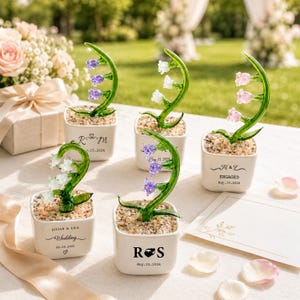 May include: Five small white ceramic pots with glass lily of the valley flowers. Each pot has a different inscription, including names, dates, and the word "Engaged". The flowers have white, purple, and pink blossoms.