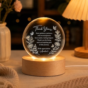 May include: A clear glass sphere resting on a round wooden base with internal LED lighting. The sphere features white etched text that reads: Thank You, Your guidance, patience, and encouragement have made a lasting impact. Thank you for being such an inspiring part of this journey. With appreciation, Amelia, surrounded by floral and butterfly designs.