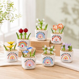 May include: Seven small white square ceramic pots containing glass flowers in various colours. Each pot features a printed rainbow, an apple, and the text Thank You for Guiding along with a name like Brown, Sophia, Oliver, or William.