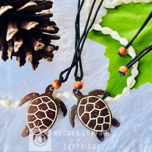 May include: Two brown turtle pendant necklaces with black cords and wooden beads. The turtle pendants have a geometric design in a lighter brown color. The background includes a pine cone, seashells, and a green leaf.