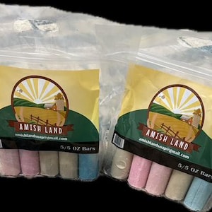 Pode incluir: Duas sacolas plásticas transparentes contendo barras de sabão coloridas. Cada sacola tem um rótulo amarelo com um logotipo circular e o texto "AMISH LAND". As barras de sabão são em tons de rosa, azul e branco. Cada barra pesa 156 gramas.