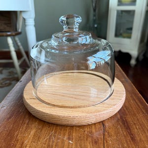 May include: A clear glass cloche with a rounded top and a small knob sits atop a round, light-colored wooden base. The cloche is designed to cover and protect food items. The wooden base has a natural wood grain.