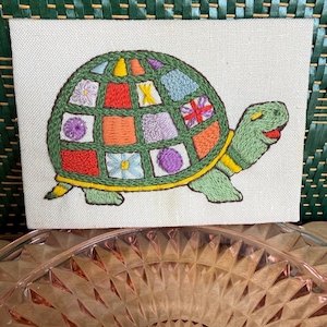 May include: Embroidered turtle art on a canvas. The turtle has a green shell with colorful patchwork squares, a yellow border, and a smiling face. The background is a woven green texture.