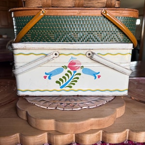 May include: A stack of vintage picnic baskets. The top basket is woven brown, the middle one is green with a wooden handle, and the bottom one is white with a floral design and metal handle.
