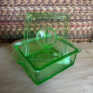 May include: A vintage, square, light green glass butter dish with a lid. The lid has a handle and a rounded, ribbed design. The dish is sitting on a wooden surface.