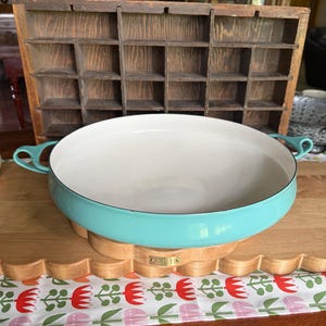 May include: A round, shallow, turquoise enameled cast iron pan with a smooth cream-colored interior and two side handles. The pan features a glossy exterior finish and is positioned in front of a wooden printer's tray with multiple small compartments.