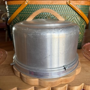 May include: A vintage aluminum cake carrier with a wooden handle and a scalloped wooden base. The carrier is cylindrical with a lid and a metal clasp. A green and brown picnic basket is in the background.