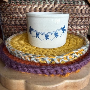 May include: A cream-colored ceramic crock with a blue heart and bow design sits atop three crocheted trivets in yellow, orange, and purple. A woven basket is in the background.